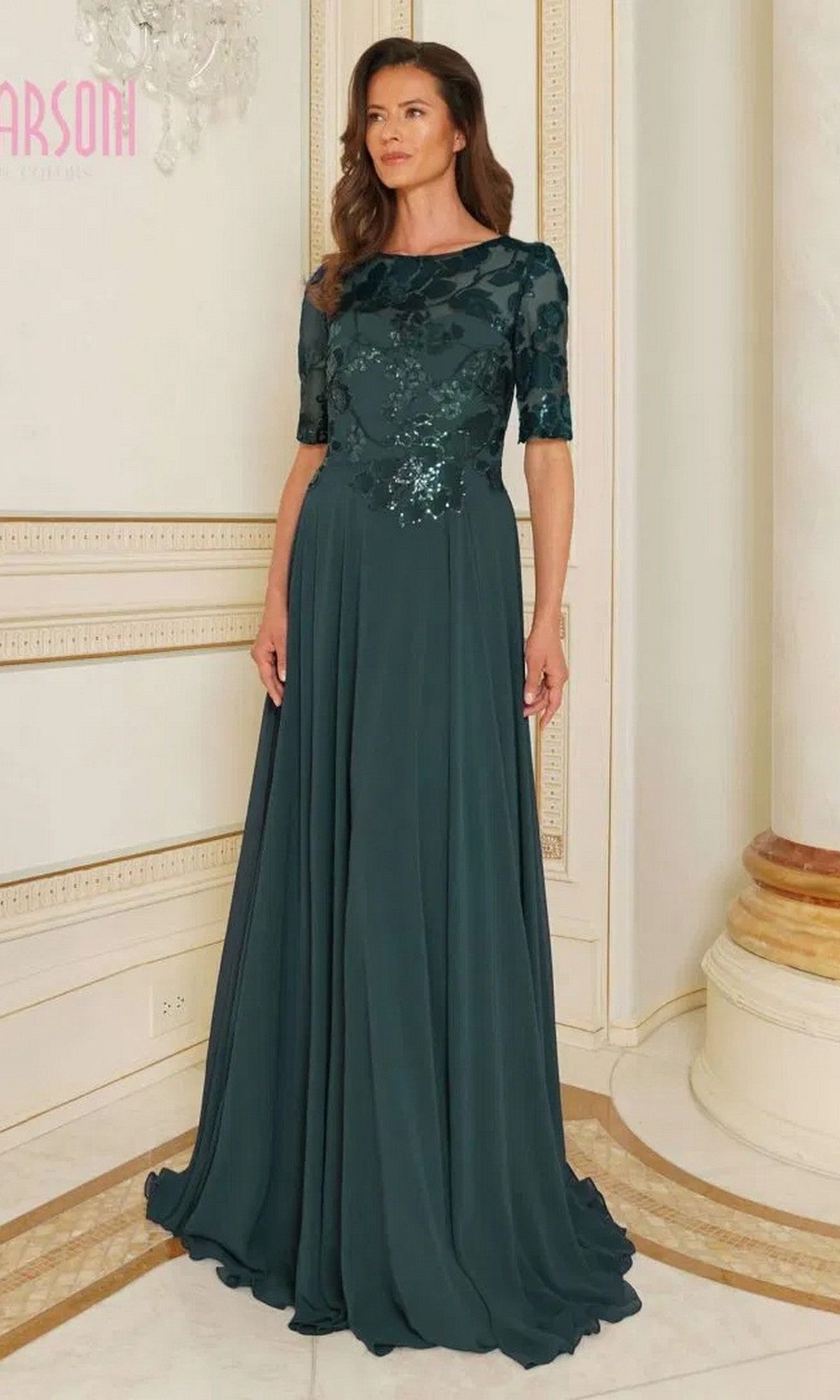 A woman stands indoors wearing the Marsoni M286 long formal dress in deep green, featuring short sleeves and a sparkling sequin lace bodice.